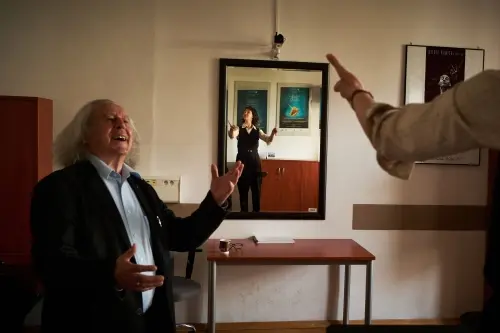 Zajęcia dyrygentury, na pierwszym planie profesor djący uwagi studence, której sylwetka odbija się w lustrze.